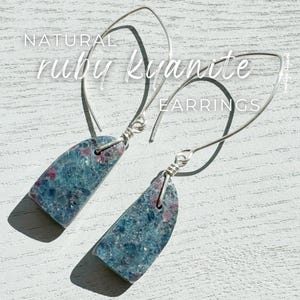 Peut inclure: Boucles d'oreilles en kyanite rubis naturelles. Ces boucles d'oreilles présentent une pierre rectangulaire bleu-vert avec des inclusions rouges, suspendue à un crochet en fil d'argent. Les boucles d'oreilles sont présentées sur une surface blanche texturée.