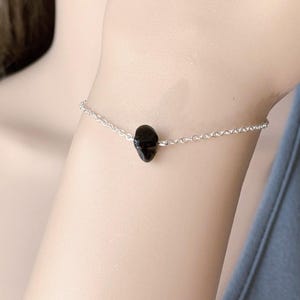 Peut inclure: Un bracelet d&eacute;licat compos&eacute; d'une fine cha&icirc;ne argent&eacute;e et d'une unique petite perle en pierre noire polie plac&eacute;e au centre. Le bracelet pr&eacute;sente une construction &agrave; maillons simples, mettant en valeur une esth&eacute;tique &eacute;pur&eacute;e avec un point focal sombre.
