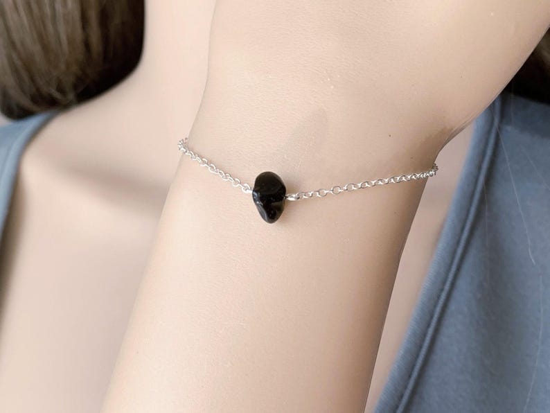 Peut inclure: Un bracelet d&eacute;licat compos&eacute; d'une fine cha&icirc;ne argent&eacute;e et d'une unique petite perle en pierre noire polie plac&eacute;e au centre. Le bracelet pr&eacute;sente une construction &agrave; maillons simples, mettant en valeur une esth&eacute;tique &eacute;pur&eacute;e avec un point focal sombre.