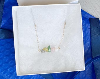 May include: A delicate gold-tone chain necklace featuring a horizontal arrangement of five small, raw, tumbled gemstone chips in shades of translucent grey, pale yellow, light green, soft pink, and clear white, resting inside a white gift box.