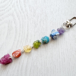 May include: A keychain featuring a series of colorful, raw gemstones in shades of pink, orange, yellow, green, blue, and purple. The stones are connected by a silver chain and attached to a silver clasp.