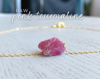 Könnte beinhalten: Eine goldene Halskette mit einem einzelnen rohen rosa Turmalin-Edelstein als zentraler Anhänger. Der Text RAW pink tourmaline ist oben im Bild zu sehen. Die Kette hat ein feines Gliederkettendesign und einen kleinen runden Verschluss.