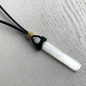 Puede incluir: Un colgante de cristal de selenita blanco en un cord&oacute;n de cuero negro con una cuenta dorada.