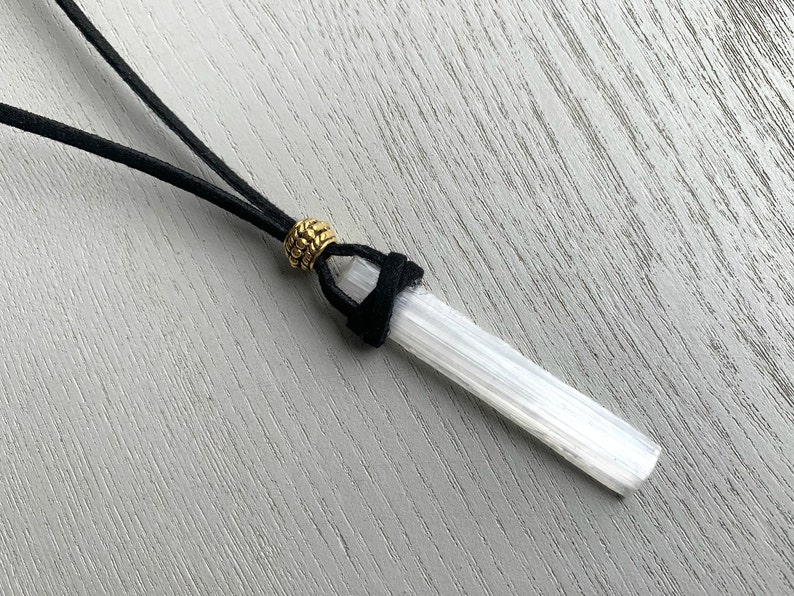 Puede incluir: Un colgante de cristal de selenita blanco en un cord&oacute;n de cuero negro con una cuenta dorada.