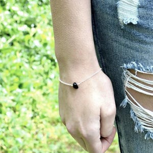 Peut inclure: Un bracelet &agrave; cha&icirc;ne fine en argent orn&eacute; d'une perle unique en pierre noire, lisse et petite, centr&eacute;e sur le poignet. Le bracelet est fix&eacute; par un fermoir simple et repose sur la peau, pr&eacute;sentant un design minimaliste avec une finition polie.