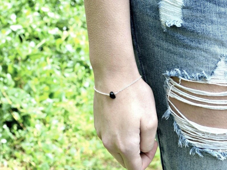 Peut inclure: Un bracelet &agrave; cha&icirc;ne fine en argent orn&eacute; d'une perle unique en pierre noire, lisse et petite, centr&eacute;e sur le poignet. Le bracelet est fix&eacute; par un fermoir simple et repose sur la peau, pr&eacute;sentant un design minimaliste avec une finition polie.