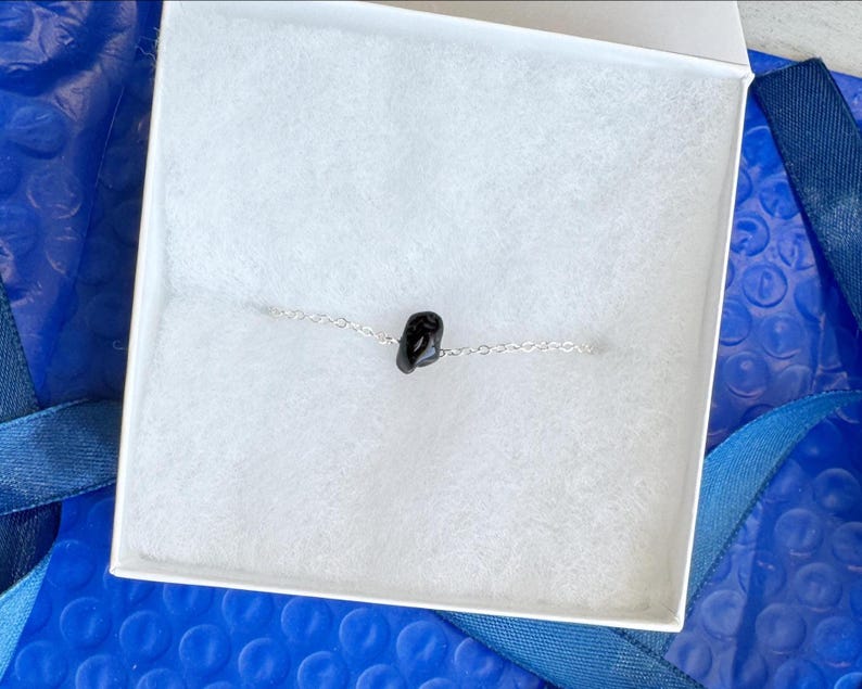 Peut inclure: Un bracelet &agrave; cha&icirc;ne argent&eacute;e d&eacute;licat comportant une seule petite perle en pierre noire polie au centre. Le bracelet est pr&eacute;sent&eacute; dans une bo&icirc;te blanche avec une doublure blanche douce et textur&eacute;e, contrastant avec la pierre sombre.