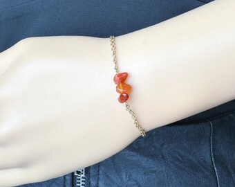 Peut inclure: Un bracelet à chaîne doré délicat orné de trois petites pierres de cornaline orange-rouge polies et de forme irrégulière, centrées sur le poignet. Le bracelet est fixé par un fermoir simple et repose sur un tissu sombre.
