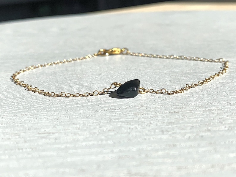 Peut inclure: Un bracelet &agrave; cha&icirc;ne dor&eacute; d&eacute;licat comportant une unique petite pierre noire lisse centr&eacute;e le long des maillons. Le bracelet comprend un petit m&eacute;canisme de fermoir en m&eacute;tal &agrave; l'arri&egrave;re pour fixer la cha&icirc;ne autour d'un poignet ou d'une cheville.