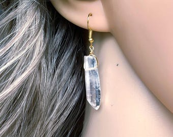 Puede incluir: Un pendiente de punta de cristal transparente y bruto suspendido de un gancho de metal dorado. El cristal está asegurado en la parte superior con alambre dorado, presentando una forma hexagonal alargada natural con inclusiones visibles.