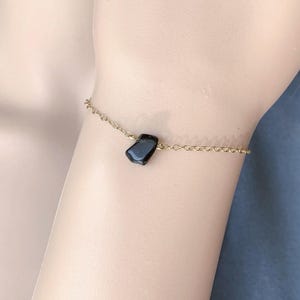 Peut inclure: Un bracelet &agrave; cha&icirc;ne fine de couleur dor&eacute;e orn&eacute; d'une unique petite pierre noire polie. Le bijou est compos&eacute; de maillons circulaires d&eacute;licats et d'un pendentif en pierre sombre &agrave; la forme irr&eacute;guli&egrave;re, centr&eacute; sur la fine cha&icirc;ne m&eacute;tallique.