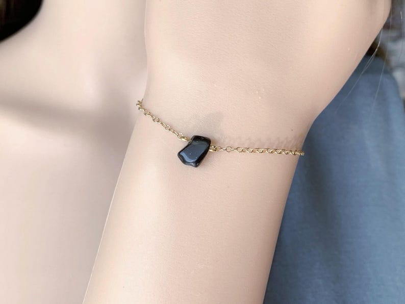 Peut inclure: Un bracelet &agrave; cha&icirc;ne fine de couleur dor&eacute;e orn&eacute; d'une unique petite pierre noire polie. Le bijou est compos&eacute; de maillons circulaires d&eacute;licats et d'un pendentif en pierre sombre &agrave; la forme irr&eacute;guli&egrave;re, centr&eacute; sur la fine cha&icirc;ne m&eacute;tallique.