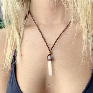 Puede incluir: Un collar de cord&oacute;n de cuero marr&oacute;n con un colgante de cristal blanco. El colgante tiene una forma rectangular larga y delgada.