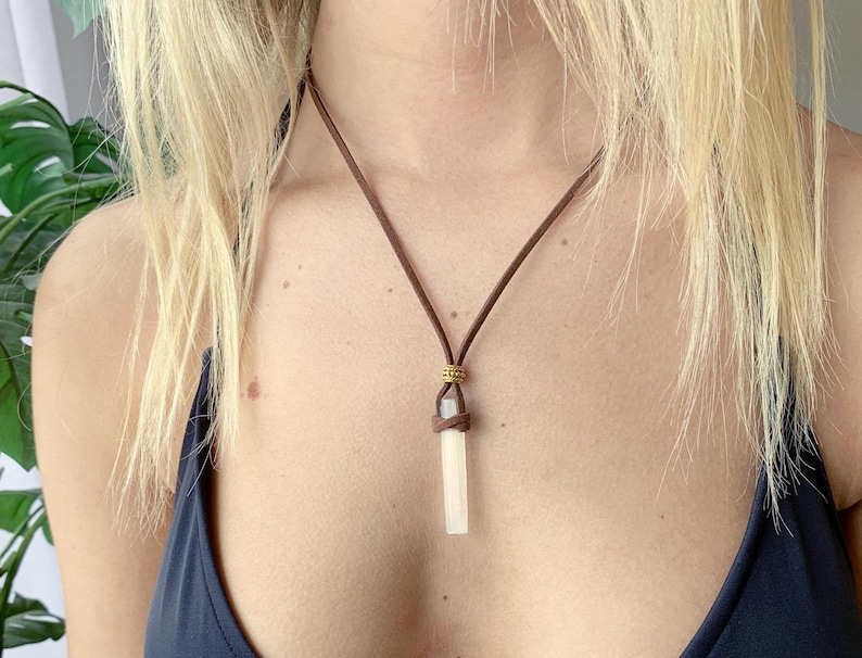 Puede incluir: Un collar de cord&oacute;n de cuero marr&oacute;n con un colgante de cristal blanco. El colgante tiene una forma rectangular larga y delgada.