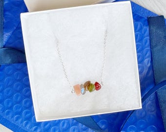 May include: A silver-toned chain necklace featuring a horizontal row of five small, polished, multi-coloured gemstone chips in shades of peach, grey, brown, green, and red, resting inside a white square gift box with white padding.