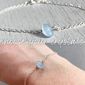 May include: A delicate silver chain bracelet featuring a single, small, polished light blue crystal stone at its center. The text "choose your crystals" is overlaid across the middle of the image, which shows the bracelet both alone and worn.