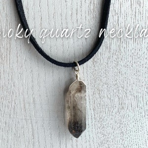 May include: A smoky quartz crystal pendant necklace on a black suede cord. The pendant is a long, pointed crystal with a dark gray color.