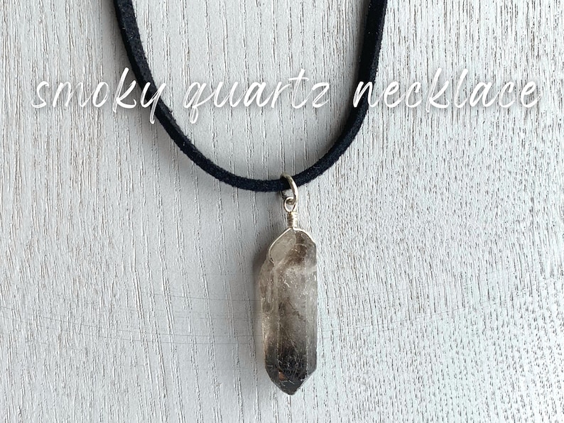 May include: A smoky quartz crystal pendant necklace on a black suede cord. The pendant is a long, pointed crystal with a dark gray color.