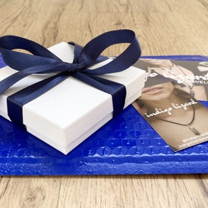 May include: A white gift box with a blue ribbon tied around it. The box is sitting on a blue bubble wrap envelope. The envelope has a business card on top that says "indigo lizard jewelry".