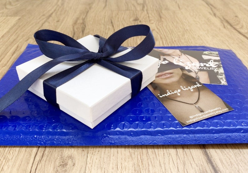 May include: A white gift box with a blue ribbon tied around it. The box is sitting on a blue bubble wrap envelope. The envelope has a business card on top that says "indigo lizard jewelry".