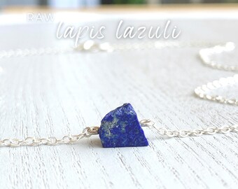 May include: A necklace featuring a single raw, deep blue lapis lazuli stone with natural gold-colored flecks, suspended on a delicate silver-toned metal chain. The text RAW lapis lazuli is visible in the upper portion of the image.