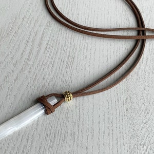 Puede incluir: Un colgante de cristal de selenita blanco en un cord&oacute;n de cuero marr&oacute;n con una cuenta dorada.