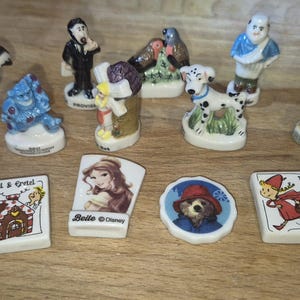 Könnte beinhalten: Eine Sammlung kleiner Keramikfiguren, darunter ein Stinktier, ein blaues Monster, ein Dalmatiner und Figuren aus Märchen. Einige Figuren tragen Text, wie "Belle @ Disney" und "Le Petit Chaperon Rouge".