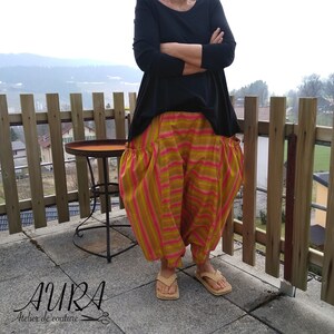 Puede incluir: Una mujer lleva una camisa negra de manga larga y pantalones a rayas con un detalle fruncido en la cintura. Los pantalones son una mezcla de rayas rosas, amarillas y naranjas. Est&aacute; de pie en un patio con una valla de madera y una mesa peque&ntilde;a en el fondo.