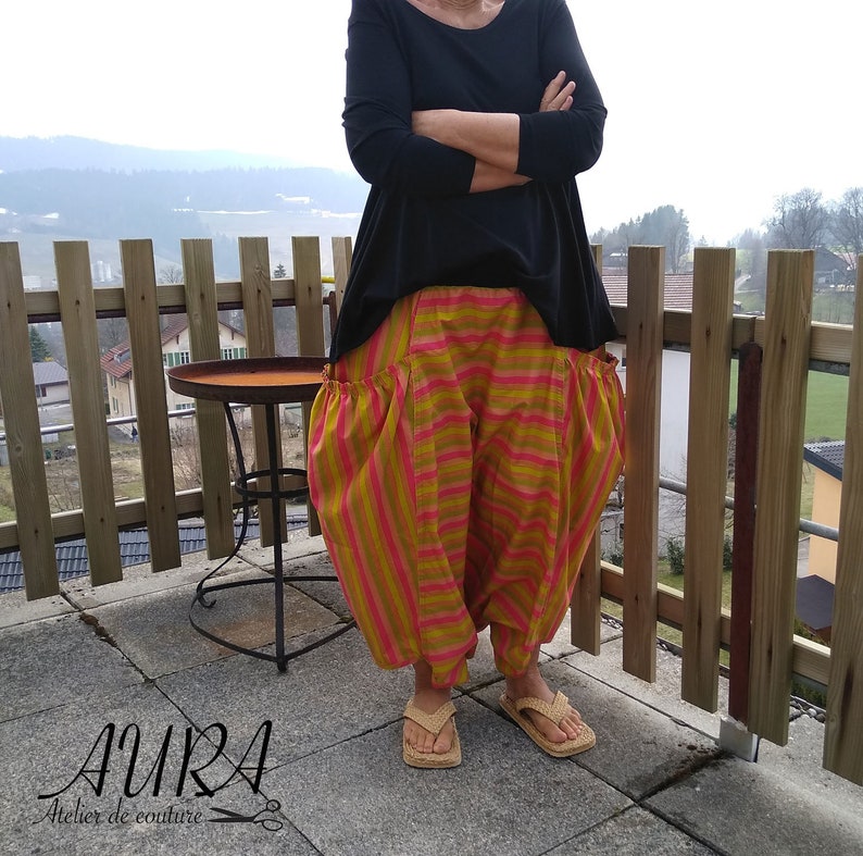 Puede incluir: Una mujer lleva una camisa negra de manga larga y pantalones a rayas con un detalle fruncido en la cintura. Los pantalones son una mezcla de rayas rosas, amarillas y naranjas. Est&aacute; de pie en un patio con una valla de madera y una mesa peque&ntilde;a en el fondo.