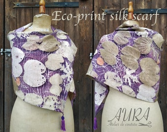 May include: Two eco-print silk scarves displayed on mannequins. The scarves feature a purple background with large, beige leaf patterns and white vein-like lines. The text "Eco-print silk scarf" is visible. Each scarf has a purple tassel.