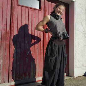 Puede incluir: Una persona joven lleva una camiseta sin mangas de rayas blancas y negras con cuello alto y una falda negra con bolsillos. Est&aacute; de pie frente a una puerta de garaje de madera roja con una ventana.