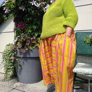 Puede incluir: Una mujer lleva un su&eacute;ter de punto verde lima y pantalones a rayas amarillas y rosas. Est&aacute; de pie frente a una planta en maceta.