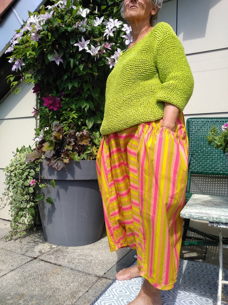 Puede incluir: Una mujer lleva un su&eacute;ter de punto verde lima y pantalones a rayas amarillas y rosas. Est&aacute; de pie frente a una planta en maceta.