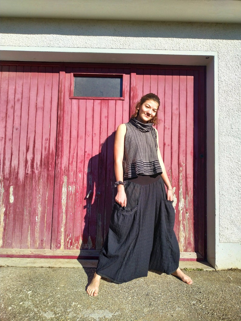 Puede incluir: Una mujer con un top sin mangas de rayas blancas y negras y pantalones negros anchos y fluidos. Est&aacute; de pie frente a una puerta de garaje de madera roja.