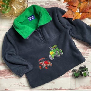 May include: A navy blue fleece pullover with a green fleece collar and a half-zip fastening. The pullover features two embroidered tractors, one green and one red, on the front. The text "Gal Gad Designs" is embroidered on a tag attached to the collar.