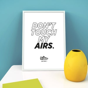 Pode incluir: Uma impressão emoldurada com o texto "DON'T TOUCH MY AIRS." e um gráfico de sapato Air Max 1. Um vaso amarelo e um bloco de notas verde estão sobre uma superfície branca. O fundo é uma parede azul clara.