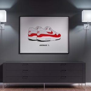 Puede incluir: Ilustración enmarcada de dos zapatillas Air Max 1 blancas, rojas y grises. Las zapatillas están representadas en una ilustración estilizada con el texto "AIRMAX 1." debajo. La obra de arte se exhibe sobre una cómoda gris oscura.