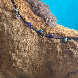 May include: A brown plush horse with a purple cord with beads and a charm attached. The beads are purple, iridescent, and gold.