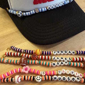 May include: A black trucker hat with a white front panel and a beaded band with the name "AUSTIN". Below are several beaded bracelets with names like "JORDAN", "NOAH", "OLIVIA", and "HUDSON", featuring sports-themed and smiley face charms.