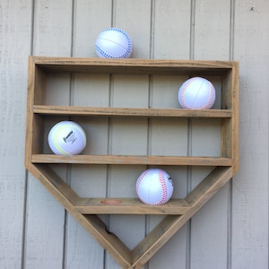 May include: A wooden baseball-shaped shelf with three shelves, holding four white baseballs with red and blue stitching. The shelf is mounted on a light beige wall.