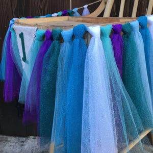 Puede incluir: Una silla decorada con una guirnalda de tul en tonos turquesa, azul, morado y blanco. La guirnalda tiene una pancarta con el n&uacute;mero "1" en ella.