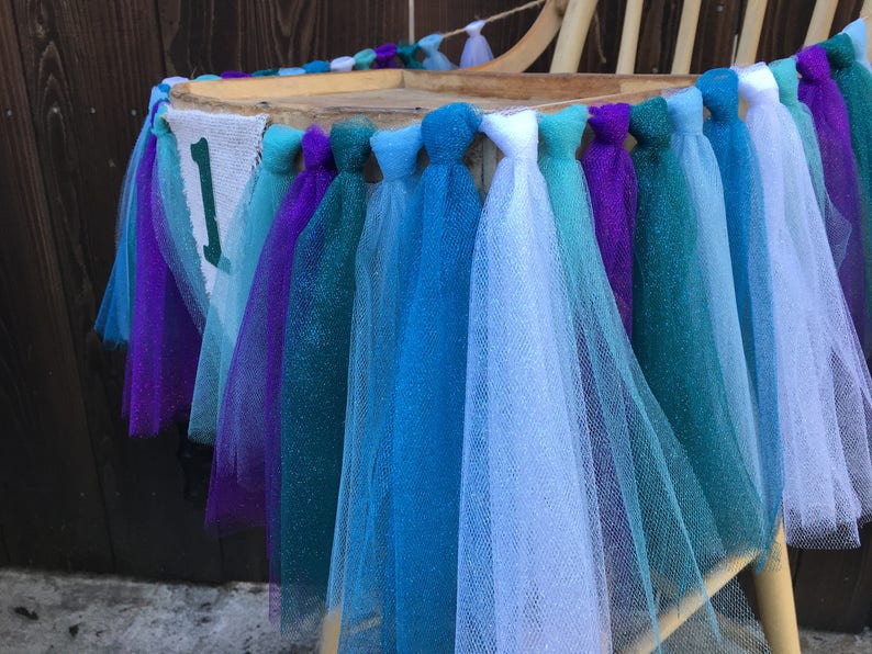 Puede incluir: Una silla decorada con una guirnalda de tul en tonos turquesa, azul, morado y blanco. La guirnalda tiene una pancarta con el n&uacute;mero "1" en ella.