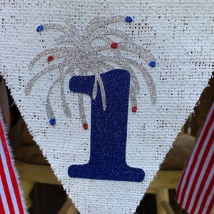 May include: A white burlap banner with a blue number one and silver glitter fireworks. The banner has red and blue glitter accents.