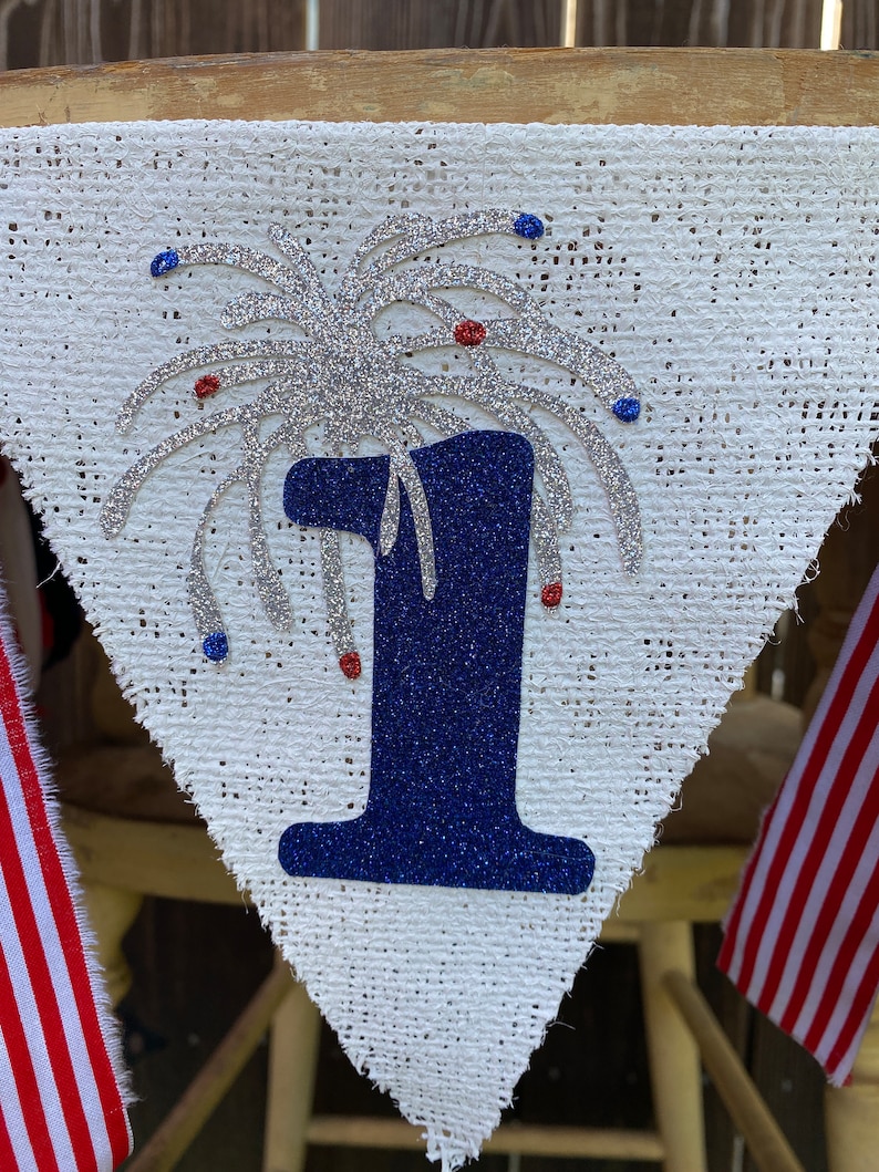 May include: A white burlap banner with a blue number one and silver glitter fireworks. The banner has red and blue glitter accents.