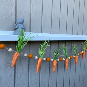 May include: A felt carrot garland with orange and white carrots and brown and white pom poms hanging from a white shelf.