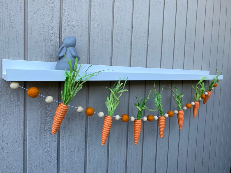 May include: A felt carrot garland with orange and white carrots and brown and white pom poms hanging from a white shelf.