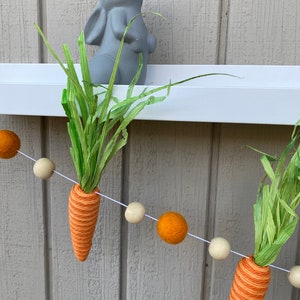 May include: A string of felt balls in orange and cream colors with a carrot and green grass hanging from it. The carrot is orange and white with green grass at the top. A gray bunny figurine is on a white shelf behind the garland.