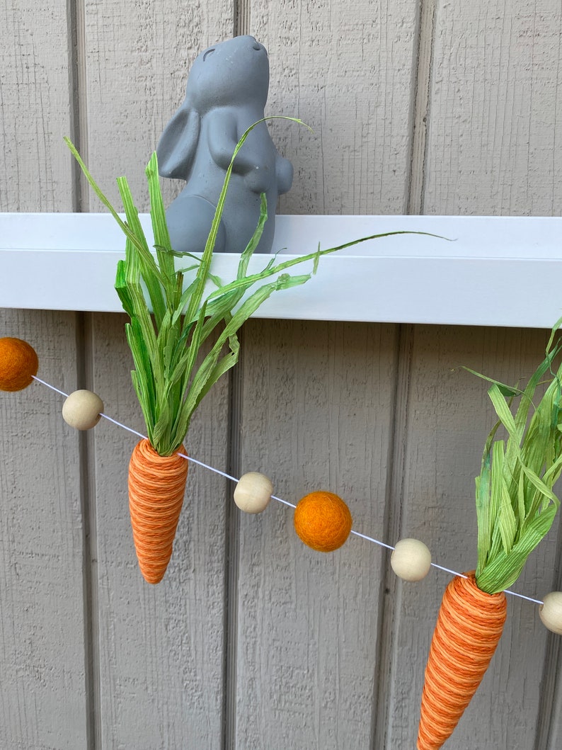 May include: A string of felt balls in orange and cream colors with a carrot and green grass hanging from it. The carrot is orange and white with green grass at the top. A gray bunny figurine is on a white shelf behind the garland.