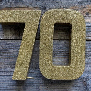 May include: Two gold glitter numbers, 7 and 0, are displayed against a wood background. The numbers are large and three-dimensional.