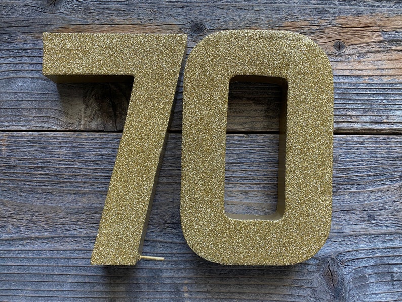 May include: Two gold glitter numbers, 7 and 0, are displayed against a wood background. The numbers are large and three-dimensional.
